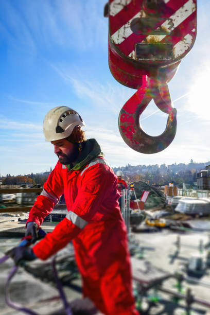 Professional manual worker attaching the load with slings to the crane, the hook is in the foreground. City and construction site in the background. Sunny day.